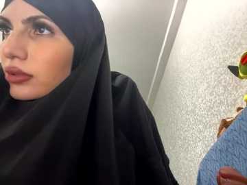 Media: Video of a woman with a light olive complexion wearing a black hijab, looking contemplative, in a room with a textured white wall and blue and yellow patterned fabric in the background.