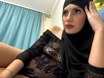 Media: Video of a light-skinned woman with brown eyes and full lips, wearing a black lace top and hijab, sitting against a teal curtain background, holding a green object.