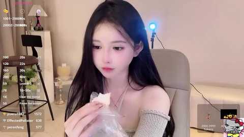 Media: Video of a young East Asian woman with long black hair, fair skin, and large eyes, wearing a beige off-shoulder sweater, sitting in a modern, beige office chair in a minimalist room.