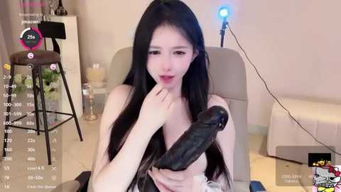 Media: A video of an East Asian woman with long black hair, fair skin, and small breasts, holding a large, erect black dildo, in a modern living room with a beige couch, a lit blue lamp, and a glass-topped table.