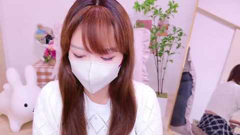 Media: Video of an East Asian woman with long brown hair and bangs, wearing a white face mask, white sweater, and white gloves, in a softly lit room with a bunny toy and a potted plant.