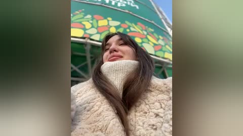 Media: Video of a woman with long brown hair wearing a beige fur coat, taken from a low angle, showing a green and yellow striped tent in the background.