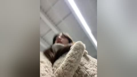 Media: A blurry video of a person in a beige, knitted sweater, lying on a beige, fuzzy blanket, with a white ceiling and fluorescent lights in the background.