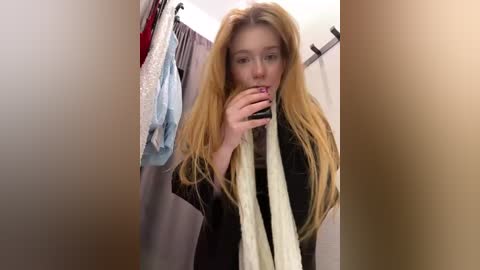 Media: A young, blonde woman with fair skin and long hair takes a selfie in a dimly lit closet, holding a beige scarf to her mouth. Clothes hang on the left and right.