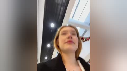 Media: Video of a blonde woman in a black blazer, standing in an indoor space with modern, white, and black architectural elements. The image is slightly blurred.