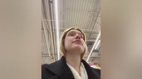 Media: Video of a blonde woman in a black coat, standing in a grocery store with fluorescent lighting and white ceiling.