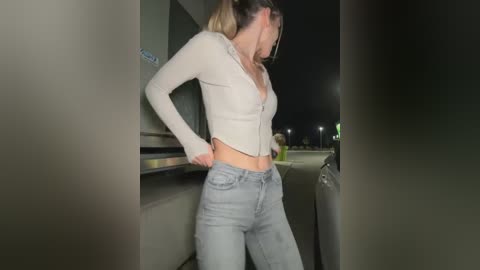 Media: Video of a young woman with light skin and blonde hair in a high ponytail, wearing a light beige, long-sleeved, cropped cardigan and light blue jeans. She stands in a dimly lit alley at night, with streetlights and distant figures visible in the background.