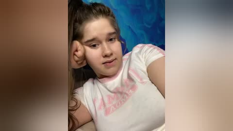 Media: Video of a young girl with light skin and long brown hair, wearing a white t-shirt with pink text, resting her head on a person's shoulder against a blue background.