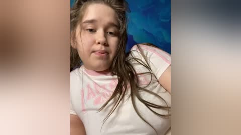 Media: Video of a young girl with light skin and wet, brown hair, wearing a white T-shirt with pink floral patterns. Background features a blue and green abstract design.