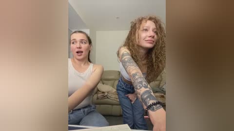 Media: Video of two women, one with light skin and straight hair, wearing a white tank top and blue jeans, and the other with curly red hair, tattoos, and a gray tank top, both looking shocked, standing in a modern living room with beige furniture.