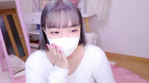 Media: A video of a young East Asian woman with straight black hair and bangs, wearing a white face mask and a white fuzzy sweater, in a cozy room with light-colored walls, a vanity table, and a pink blanket.