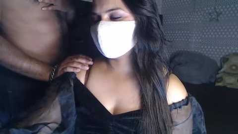 Media: Video of a topless woman with long black hair, wearing a face mask, holding a man's hand. She has a dark complexion and is in a dimly lit room with grey patterned wallpaper.