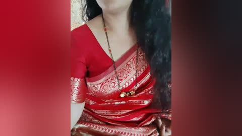 Media: Video of a woman in a vibrant red sari with gold embroidery, wearing a black necklace, against a gradient red background. Her long, dark hair cascades over her shoulders.