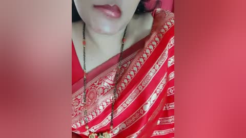 Media: A close-up video of a woman's face and neck, wearing a red and gold-patterned saree, with a black necklace featuring a pendant. Her lips are slightly parted, revealing a glossy pink lipstick. The background is blurred in a warm red hue.