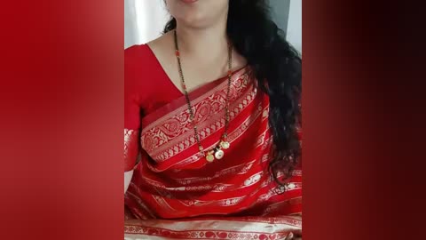 Media: Video of a woman in a red sari with intricate gold embroidery, wearing a gold necklace with small pendants, against a blurred red background.