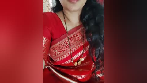 Media: Video of a woman in a red sari with intricate gold embroidery, wearing a black beaded necklace, with long black hair, against a red and black gradient background.