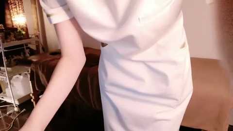 Media: Video of a slender, light-skinned woman in a white nurse uniform, standing in a dimly lit room with a bed, lamp, and cluttered desk in the background.