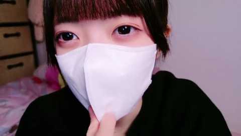 Media: A close-up video of an Asian woman with short black hair, wearing a white face mask, black top, and looking directly at the camera. Background includes a wooden dresser and a bed with colorful bedding.