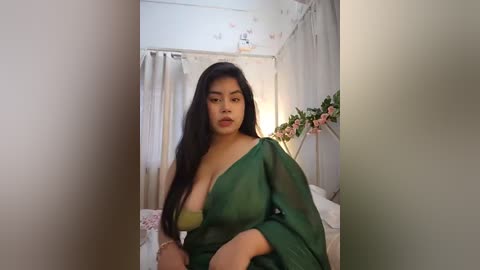 Media: Video of a young woman with long black hair, wearing a green, sheer robe, revealing a green bra, standing in a softly lit bedroom with white walls, curtains, and floral decorations.