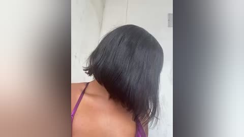 Media: A video of a woman with a sleek, straight black bob haircut, wearing a purple spaghetti-strap top. The background features a plain white wall with a small, dark rectangular object.