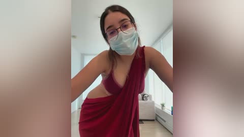 Media: Video of a young woman with light skin and brown hair, wearing a red saree, blue surgical mask, and glasses, standing in a bright, modern living room with white walls and large windows.