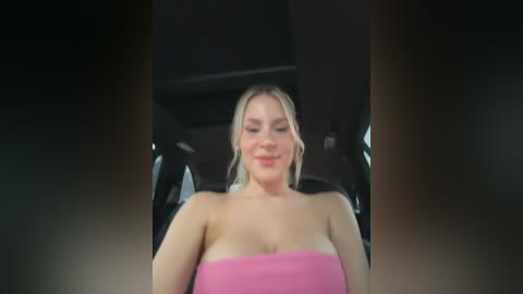 Media: Video of a smiling, blonde woman with fair skin, wearing a strapless pink dress, sitting in a dimly lit car interior.