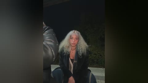 Media: Video of a blonde woman with wavy hair, wearing a black leather jacket and jeans, sitting on a concrete floor against a dark background.