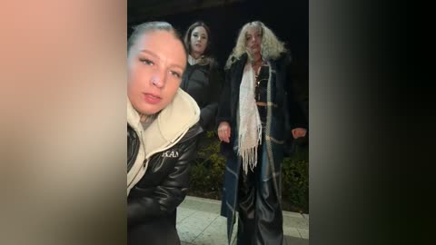 Media: A video captures three women in winter attire, including a blonde in a black leather jacket and white scarf, a brunette in a dark coat, and a woman with shoulder-length hair in a blue coat. The background features a garden and a dark, dimly lit environment.