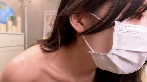 Media: Video of a woman with shoulder-length brown hair, wearing a white surgical mask, in a blurred background featuring white furniture and a blue object.