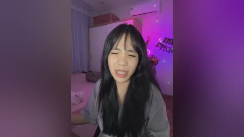 Media: A video of an Asian woman with long black hair, wearing a grey shirt, smiling, in a dimly lit room with purple lighting, white walls, and a bed in the background.