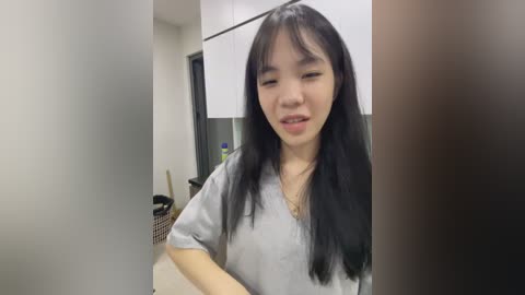 Media: Video of an East Asian woman with long black hair, wearing a light gray V-neck shirt, standing in a modern kitchen with white cabinets, a basket on the floor, and blurred background.