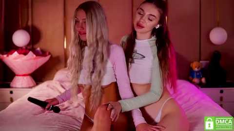 Media: Video of two young women with long blonde hair, wearing crop tops, sitting on a bed, holding a microphone.