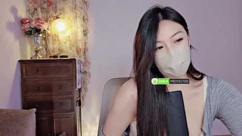 Media: Video of an Asian woman with long black hair, wearing a gray top, face mask, and black smartphone. Background includes a wooden dresser with a lamp and flowers.