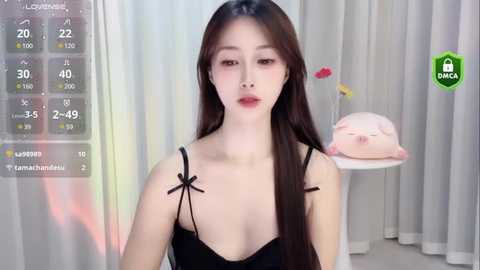 Media: A video of a young East Asian woman with long brown hair, wearing a black spaghetti strap top, standing in a softly lit room with a pink pillow and white curtains.