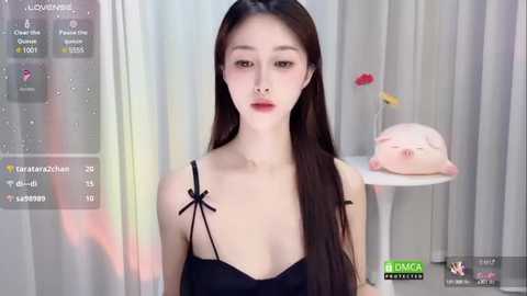 Media: Video of a young East Asian woman with long black hair and fair skin, wearing a black spaghetti-strap top, standing in a modern, minimalistic room with light-colored curtains.