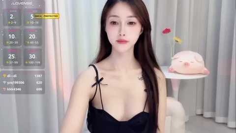 Media: A video of an Asian woman with long black hair, fair skin, and a slender physique, wearing a black camisole. Background includes a virtual reality display, a pink piggy bank, and floral curtains.