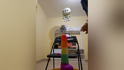 Video of a small, dimly lit room with yellow walls, featuring a black chair, a toy astronaut on a shelf, and a colorful, rainbow-striped dildo on the floor.