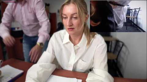 Media: Video of a blonde woman in a white blouse, sitting at a table, with blurred background of two people.