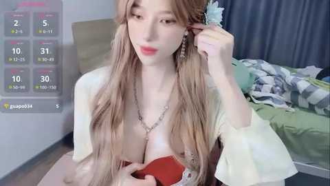 Media: Video of a young Asian woman with long, wavy blonde hair, wearing a revealing white top, adjusting her hair. Background shows a messy bedroom with a green bedspread.