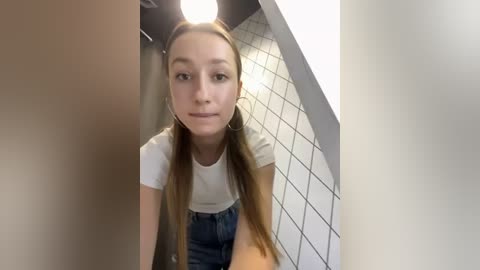 Media: Video of a young woman with long, straight brown hair in pigtails, wearing a white t-shirt and blue jeans, standing in a tiled restroom. She has a neutral expression.