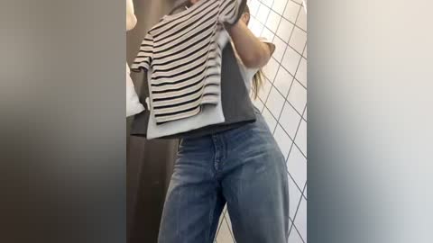 Media: Video of a person wearing a striped t-shirt, grey top, and blue jeans, lifting a shirt over their head in a changing room with white tiled walls and dark curtains.