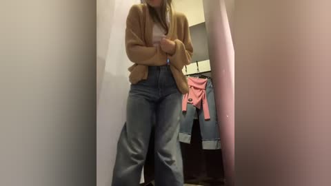 Media: A video shows a young woman with long hair in a tan cardigan and high-waisted jeans, standing in a narrow hallway. Pink and red clothes hang behind her.