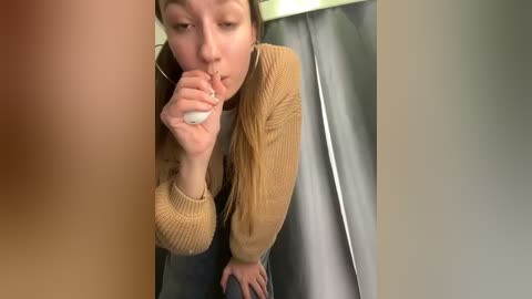 Media: A video of a young woman with light skin and long, light brown hair, wearing a tan sweater, bent over a urinal, holding a white sanitary pad. Background shows a gray curtain and beige wall.