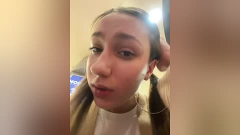 Media: Video of a young woman with light skin, brown hair tied back, wearing a beige jacket over a white shirt, making a pouty face, taken in a dimly lit room with blurred background.