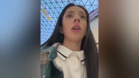 Media: A video of a young woman with long, dark hair and fair skin, wearing a white blouse and a teal blazer, standing in a modern, glass-roofed atrium with geometric patterns.