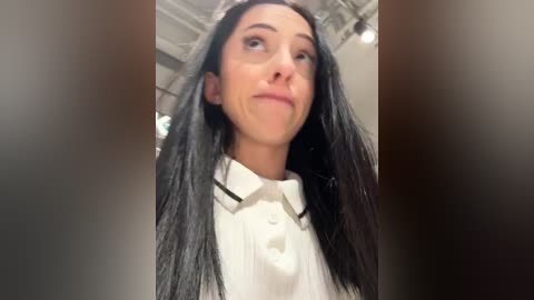 Media: Video of a young woman with long, straight black hair, pale skin, and a confident expression. She wears a white collared shirt, indoors with a blurred background.