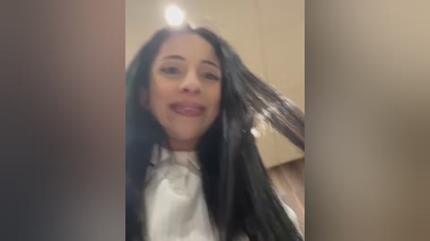 Media: Video of a smiling woman with long, straight black hair, wearing a white shirt, taken from an angled view, indoors with a blurred background.