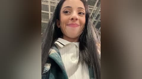 Media: Video of a young woman with long black hair, wearing a white collared shirt under a green plaid sweater, smiling in a modern indoor setting with a grid ceiling.