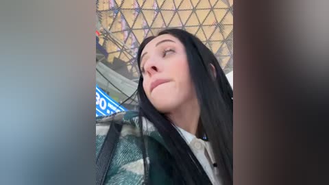 Media: Video of a young woman with long black hair, fair skin, and closed eyes, wearing a green and white patterned jacket. Background features a colorful, geometrically-patterned mural.