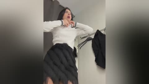 Media: Video of a woman in a white sweater and black feathered skirt, looking shocked, standing in a changing room with a black jacket on a hanger.